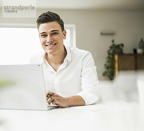 Lächelnde männliche Fachkraft  die einen Laptop benutzt  während sie an einem Tisch im Heimbüro sitzt
