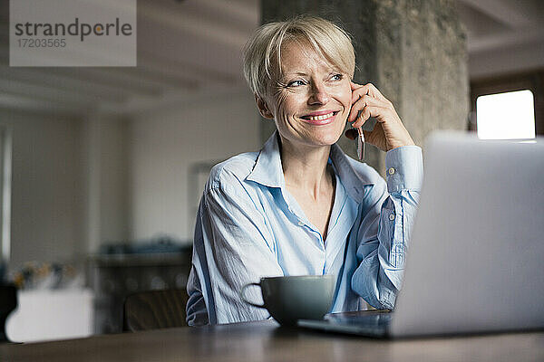 Lächelnde Geschäftsfrau mit Laptop und Kaffeetasse  die wegschaut  während sie am Schreibtisch im Heimbüro mit dem Handy telefoniert