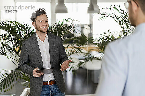 Lächelnder Geschäftsmann mit digitalem Tablet  der mit einem männlichen Kollegen im Büro diskutiert
