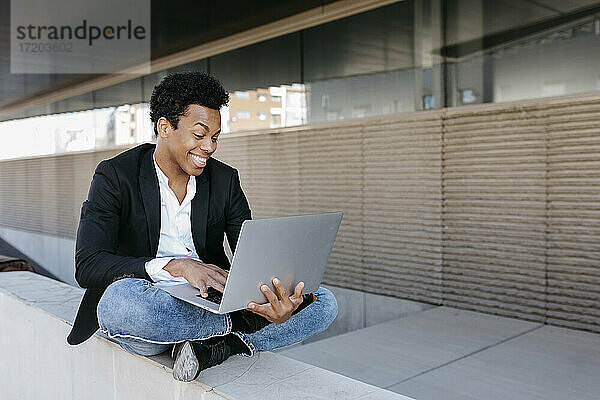 Lächelnder Geschäftsmann  der einen Laptop benutzt  während er im Schneidersitz auf einer Stützmauer sitzt