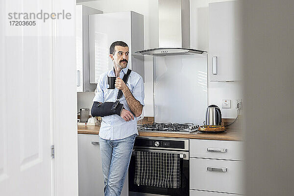 Mann trägt Armschlinge beim Teetrinken in der Küche zu Hause
