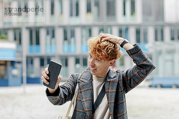 Rothaariger Mann mit Hand im Haar  der ein Selfie mit seinem Smartphone im Freien macht