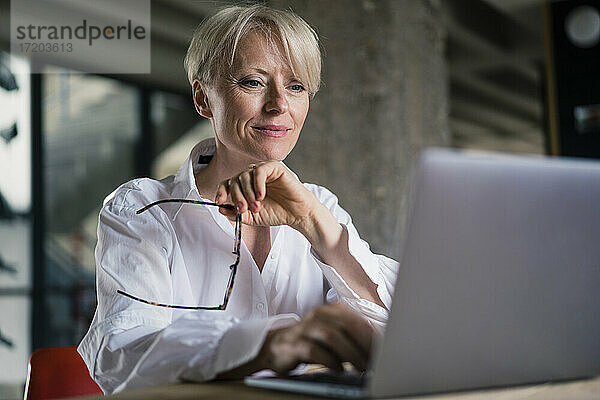Lächelnde Geschäftsfrau mit Brille bei der Arbeit am Laptop im Büro zu Hause