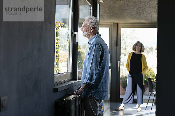Älterer Mann schaut durch ein Fenster  während er mit einer Frau im Hintergrund zu Hause steht