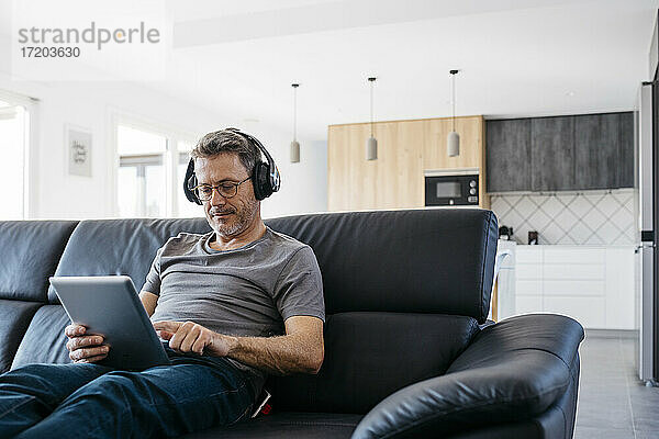 Älterer Mann mit Kopfhörern  der ein digitales Tablet benutzt  während er zu Hause auf dem Sofa sitzt