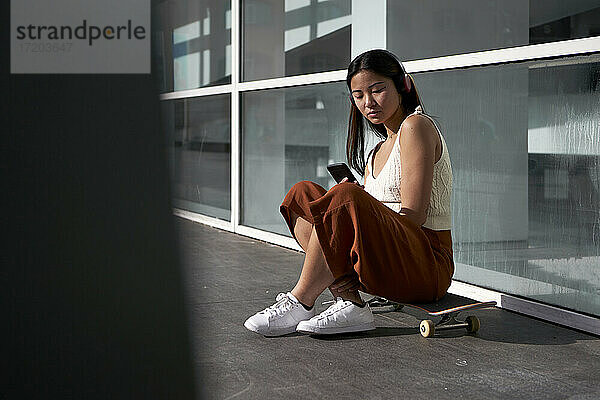 Asiatische Frau  die ein Smartphone benutzt  während sie auf einem Skateboard sitzt  an einem sonnigen Tag