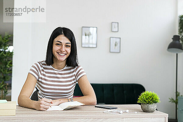 Fröhliche Frau mit Notizbuch und Stift am Wohnzimmertisch sitzend