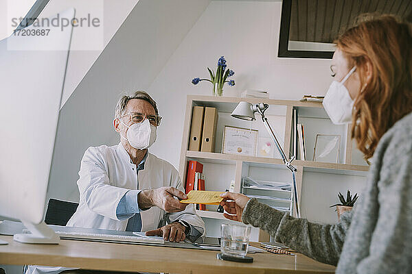 Männlicher Arzt mit Gesichtsschutzmaske  der einem Patienten im Büro sitzend einen Impfpass aushändigt