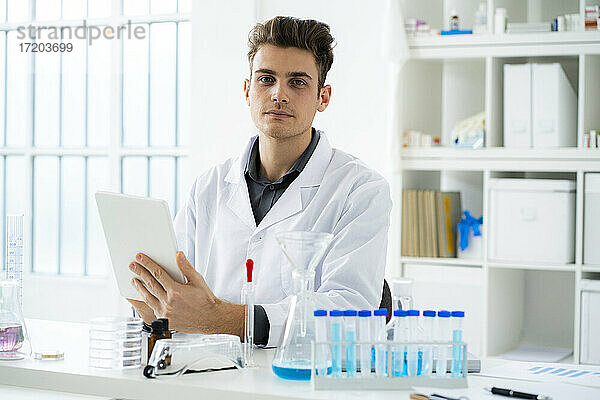 Junger männlicher Wissenschaftler  der ein digitales Tablet hält  während er am Schreibtisch in einem Labor sitzt