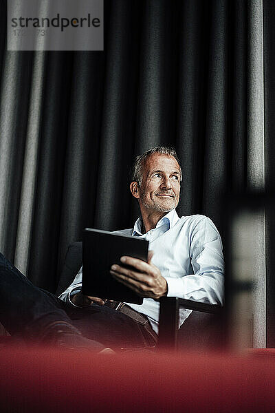 Lächelnder männlicher Geschäftsmann  der wegschaut und ein digitales Tablet auf einem Sessel in einer Büro-Cafeteria hält
