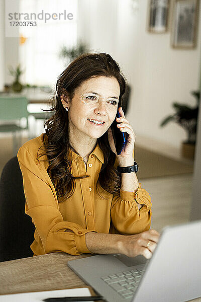 Lächelnde Geschäftsfrau mit Laptop  die auf einem Smartphone spricht  während sie am Schreibtisch im Heimbüro sitzt