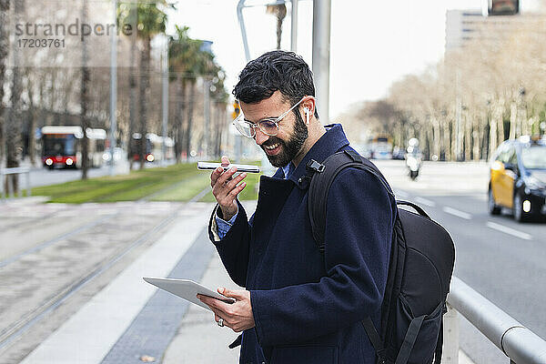Lächelnder Geschäftsmann mit digitalem Tablet  der mit seinem Smartphone über einen Lautsprecher am Bahnhof spricht