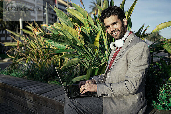 Lächelnder männlicher Unternehmer  der einen Laptop benutzt  während er auf einer Stützmauer sitzt