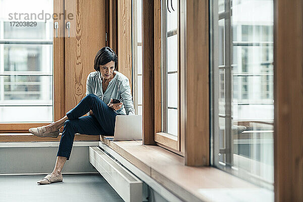 Geschäftsfrau arbeitet am Laptop  während sie im Büro am Fenster sitzt
