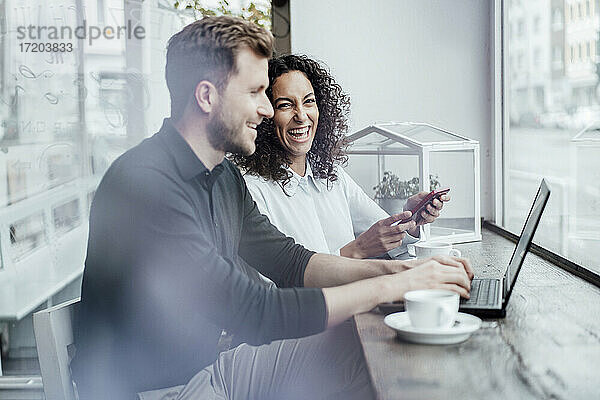 Lächelnde Geschäftsfrau mit Mobiltelefon neben einem Kollegen  der in einem Café am Laptop arbeitet