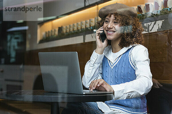 Lächelndes Teenager-Mädchen mit Baskenmütze  das mit einem Laptop in einem Café sitzt und mit einem Mobiltelefon spricht