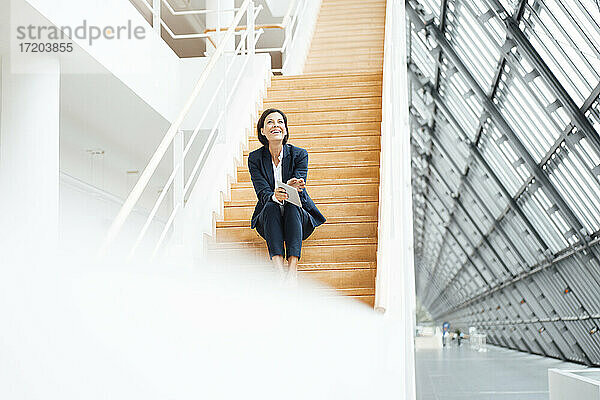 Geschäftsfrau mit digitalem Tablet  die lachend auf einer Treppe im Korridor sitzt