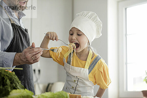 Mädchen mit Schürze und Kochmütze isst Nudeln mit Vater in der Küche zu Hause