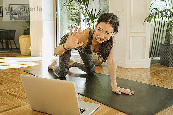 Lächelnde  schöne Lehrerin  die während eines Online-Yoga-Kurses über einen Laptop winkt