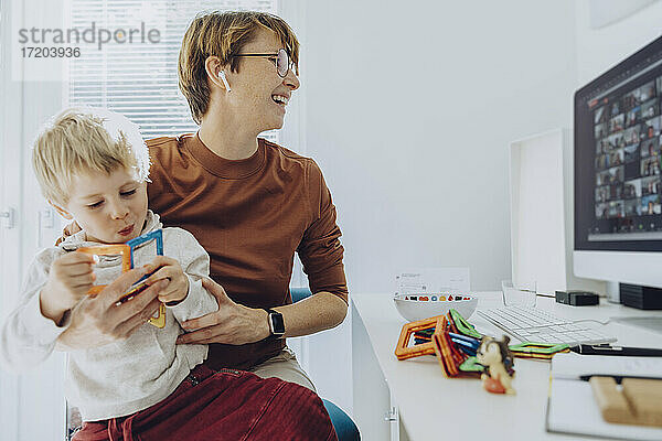 Mid adult businesswoman working while son sitting on her lap playing with toys at home office