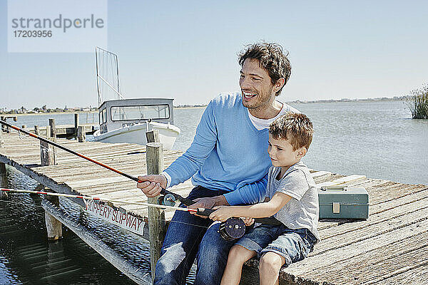 Fröhlicher Vater beim Angeln mit seinem Sohn auf dem Steg sitzend