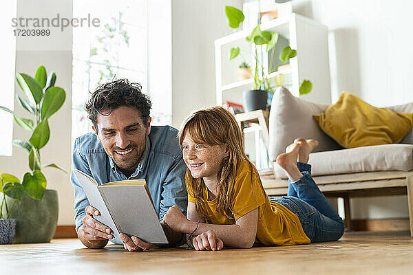 Lächelnder Vater und Tochter lesen gemeinsam ein Buch  während sie zu Hause auf dem Boden liegen