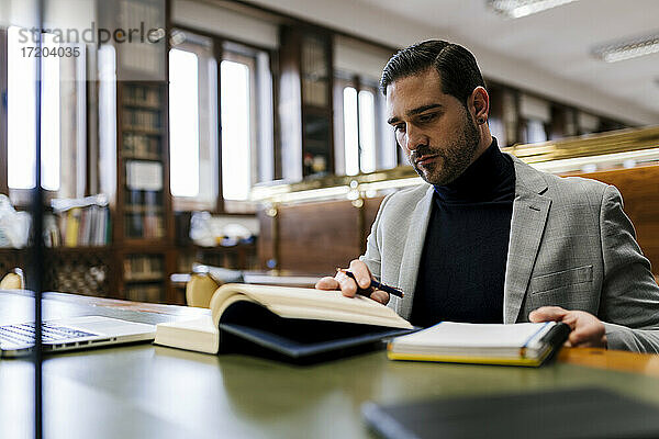 Geschäftsmann mit Laptop  der ein Buch liest  während er in einer Bibliothek arbeitet