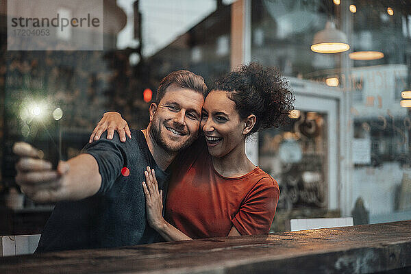 Fröhliches Paar nimmt Selfie durch Handy beim Sitzen im Café