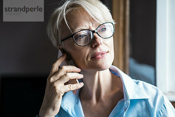 Geschäftsfrau mit Brille  die wegschaut  während sie im Büro zu Hause mit dem Handy telefoniert