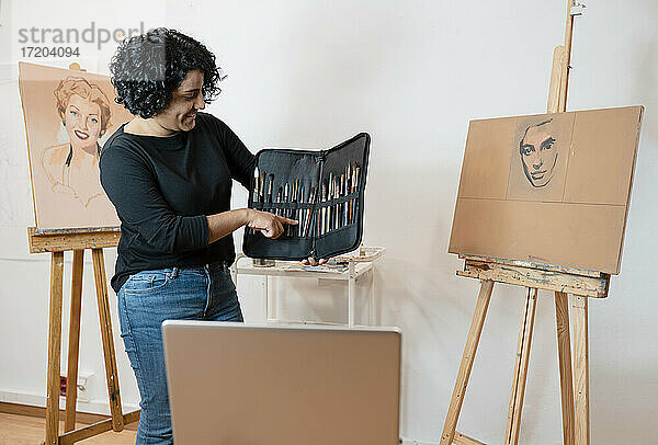 Lächelnde Malerin inmitten von Staffeleien stehend  während sie auf einen Pinsel im Kunststudio zeigt