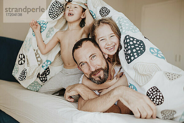 Fröhliche Tochter mit Vater  während der Bruder unter der Decke auf dem Bett zu Hause wegschaut