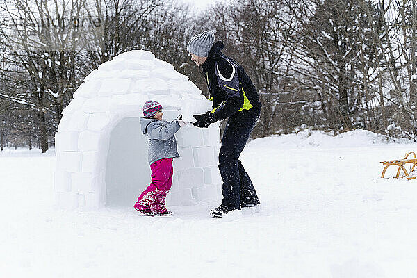 Mädchen mit Hilfe des Vaters  der einen Schneeblock trägt  um im Park ein Iglu zu bauen