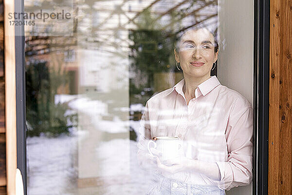 Lächelnde Unternehmerin mit Kaffeetasse  die durch ein Glasfenster wegschaut