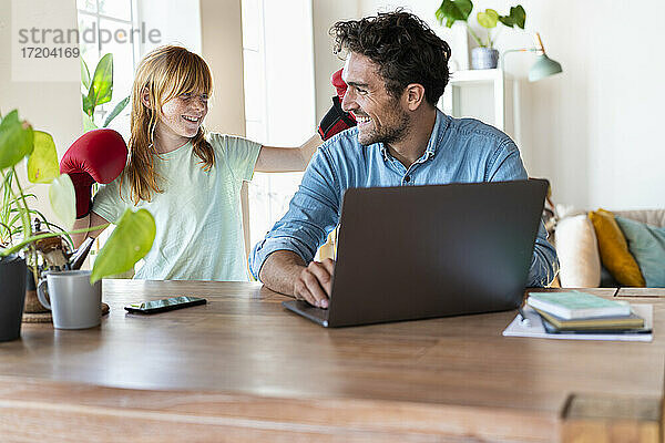 Verspieltes rothaariges Mädchen hat Spaß mit Vater  der zu Hause am Laptop arbeitet