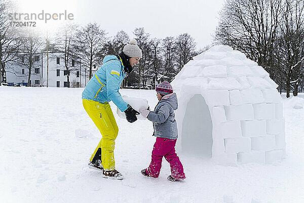 Mädchen mit Hilfe der Mutter  die einen Schneeblock trägt  um im Park ein Iglu zu bauen