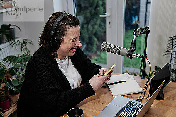 Lächelnde Frau  die während einer Podcast-Sitzung zu Hause ihr Smartphone neben dem Laptop am Schreibtisch benutzt