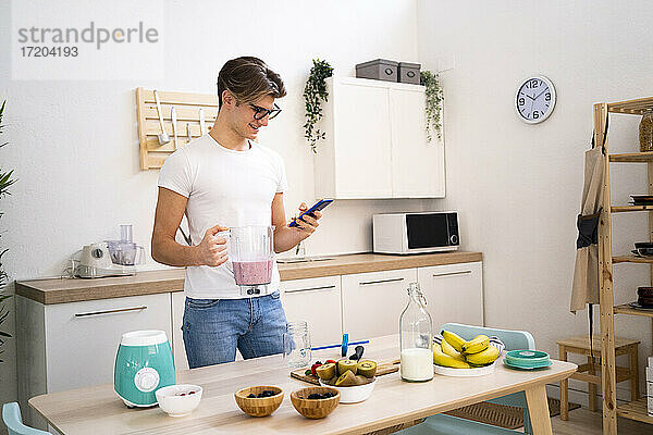 Junger Mann hält einen Smoothie-Mixer in der Hand und benutzt sein Smartphone in der Küche