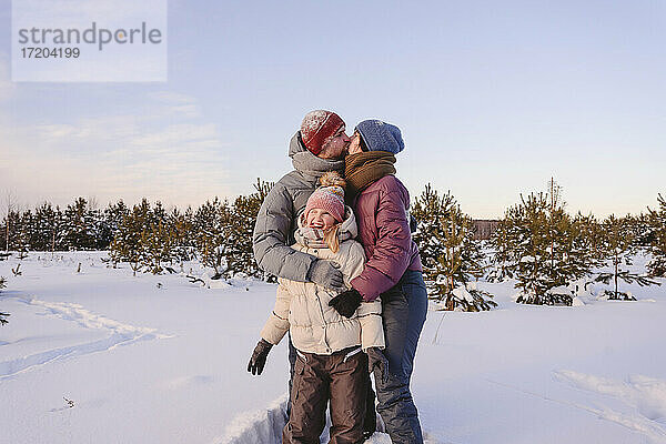 Küssende Eltern  die ihre süße Tochter im Winter im Schnee umarmen