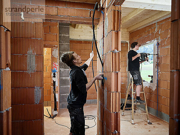 Männlicher Elektriker  der Kabel hält  während ein Kollege auf der Baustelle eine Wand anbohrt