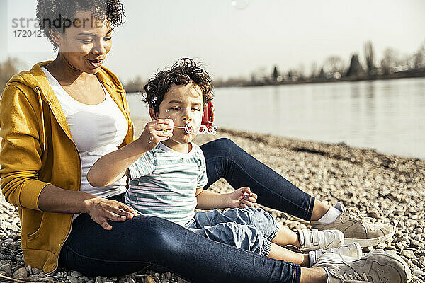 Junge bläst in den Seifenblasenstab  während er mit einer Frau auf einem Kieselstein sitzt
