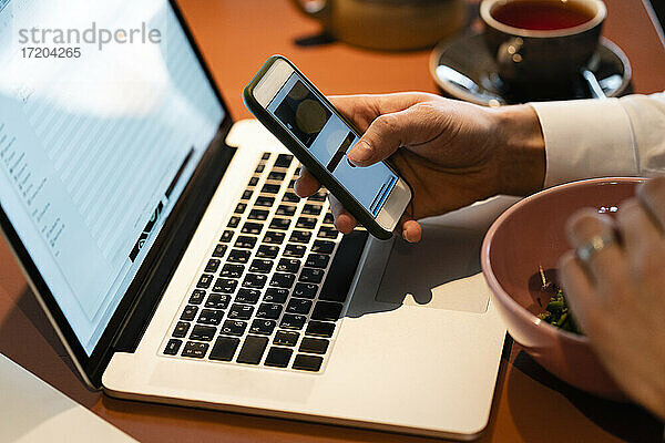 Geschäftsmann  der ein Smartphone über einem Laptop benutzt  während er in einem Café sitzt