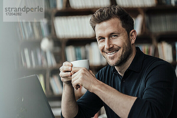 Lächelnder Geschäftsmann trinkt Kaffee und sitzt mit einem Laptop in einem Café