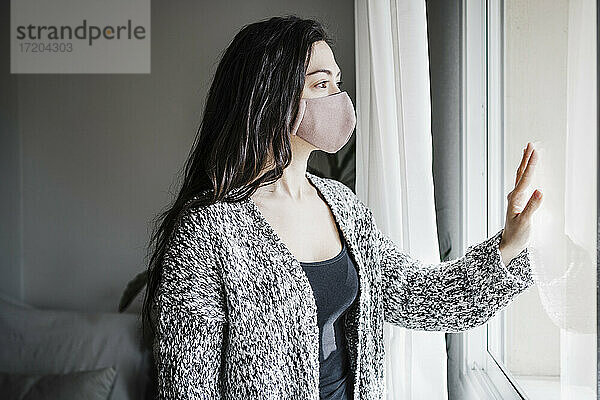 Frau mit Gesichtsschutzmaske  die durch das Fenster schaut  während sie im Wohnzimmer ihres Hauses steht  während COVID-19