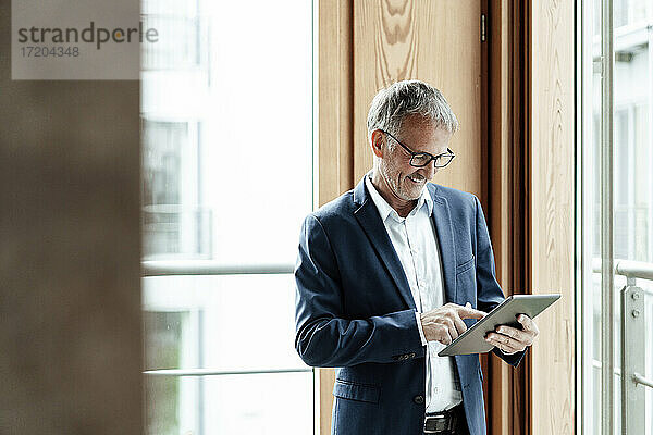 Lächelnder Geschäftsmann  der ein digitales Tablet benutzt  während er vor einem Glasfenster steht