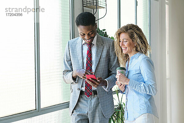 Männlicher Unternehmer  der ein Smartphone benutzt und neben einer lächelnden Kollegin mit Kaffeetasse steht