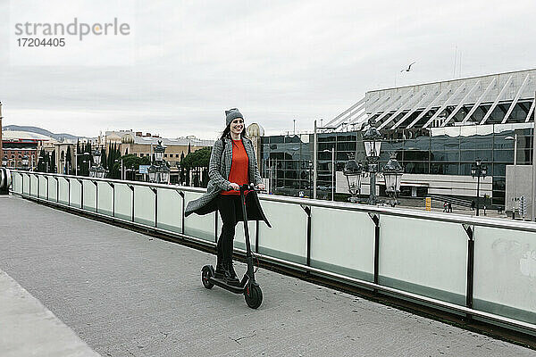 Glückliche Frau fährt elektrischen Roller auf dem Gehweg in der Nähe des Geländers in der Stadt