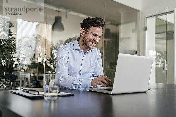 Lächelnder Geschäftsmann mit Laptop am Schreibtisch im Büro