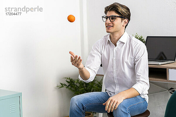 Lächelnder junger Mann  der eine Orange wirft  während er zu Hause im Wohnzimmer sitzt