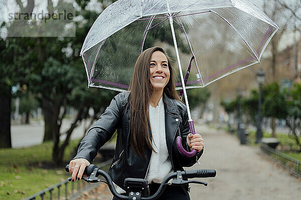 Lächelnde Frau mit Elektrofahrrad und Regenschirm auf der Straße in der Stadt