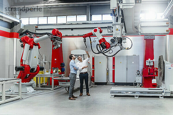 Männliche Ingenieure diskutieren über einen Roboter-Schweißbrenner  während sie an einer Maschine in einer Fabrik stehen
